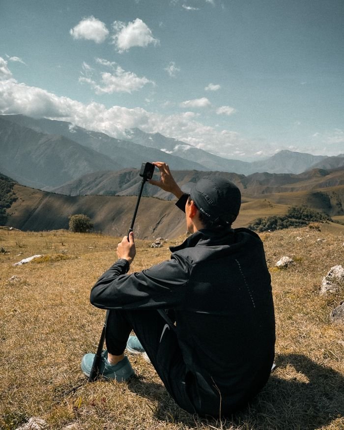 services-04 A man in active wear takes a selfie with a selfie stick on a mountain top, enjoying the scenic view.
