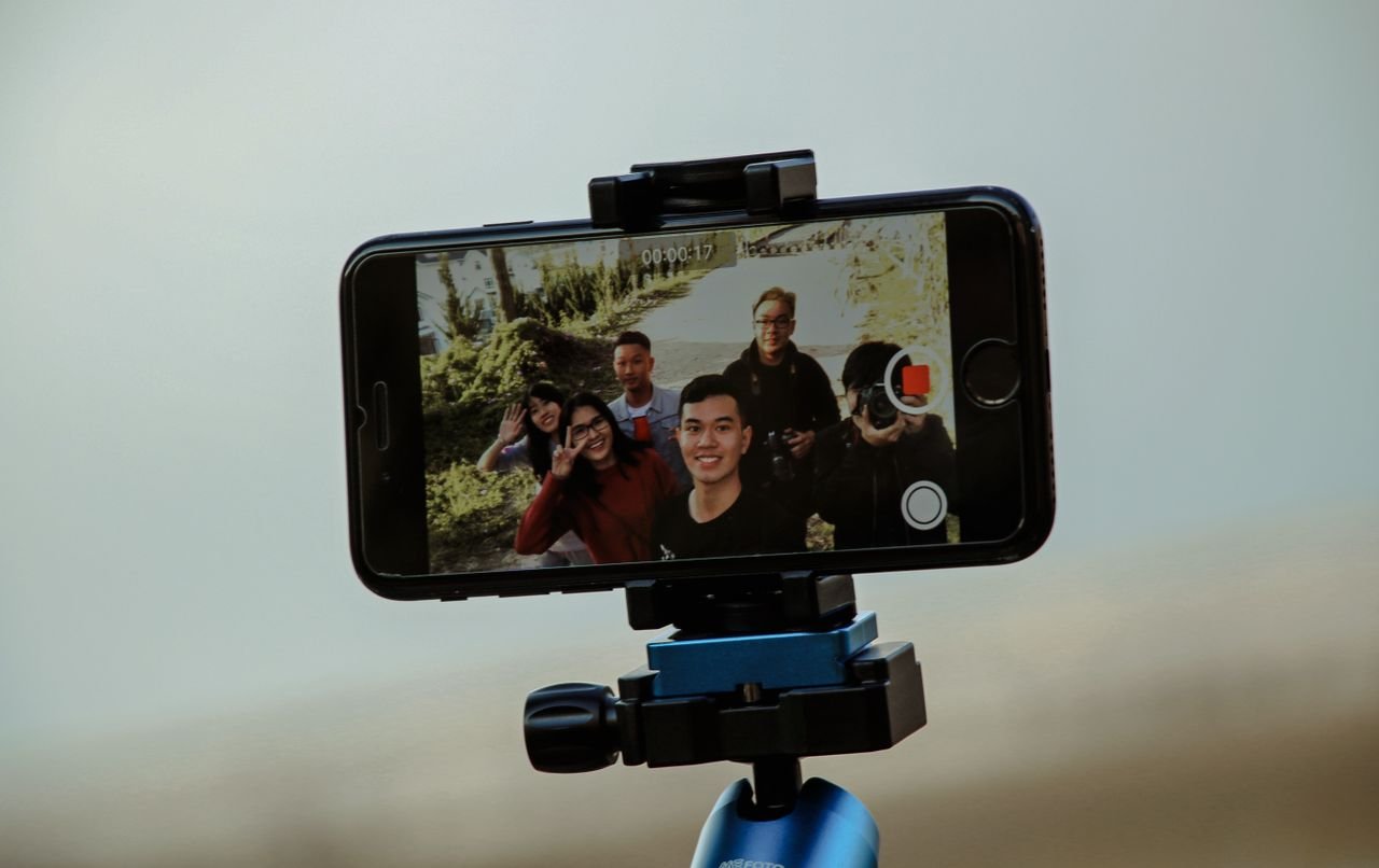 services-02 A group of friends enjoying video recording with a smartphone mounted on a tripod outdoors, capturing candid moments.